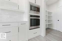 White shaker-style cabinetry with matte black hardware, stainless steel built-in microwave and oven, white subway tile backsplash, light wood-finish flooring, and a walk-in pantry with built-in shelving - 