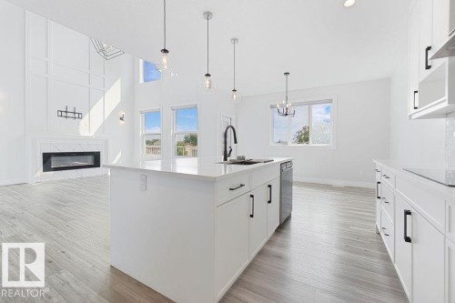Open-concept living space with wood-finish flooring, a kitchen island with pendant lighting, white cabinetry, and a fireplace with a marble-style surround - 9331 181 Avenue Nw, Edmonton, AB - Indoor Photo Showing Kitchen With Upgraded Kitchen