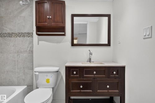 Full bathroom featuring a dark wood-finish vanity with a light-toned countertop, a matching wood-finish wall cabinet, a framed mirror, and tiled shower surround with an accent strip - 1315 Westerra Drive, Stony Plain, AB - Indoor Photo Showing Bathroom