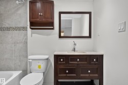 Full bathroom featuring a dark wood-finish vanity with a light-toned countertop, a matching wood-finish wall cabinet, a framed mirror, and tiled shower surround with an accent strip - 
