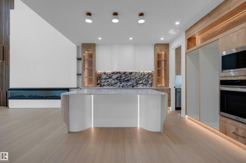 Modern kitchen featuring a waterfall island with integrated lighting, a full-height marble-patterned backsplash, light wood-finish cabinetry, stainless steel appliances, and a built-in fireplace - 2408 1 Avenue Sw, Edmonton, AB - Indoor Photo Showing Kitchen