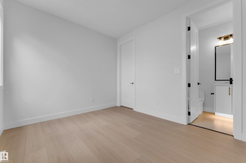 Light wood-finish flooring with white walls and trim, featuring a white solid core door with black hardware, and an additional room with a white vanity and black framed mirror - 2408 1 Avenue Sw, Edmonton, AB - Indoor Photo Showing Other Room