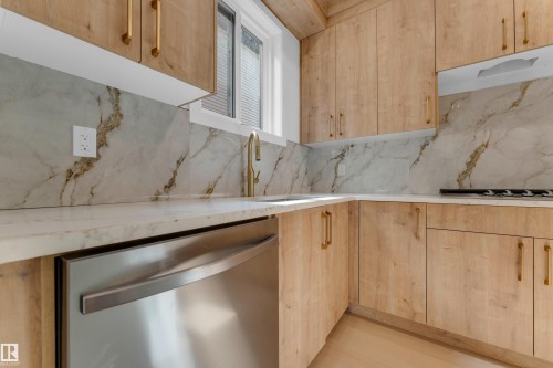 Light wood-finish cabinetry with gold-tone hardware, white and gold veined countertop, matching backsplash, stainless steel dishwasher, and a gold-tone faucet - 2408 1 Avenue Sw, Edmonton, AB - Indoor Photo Showing Kitchen
