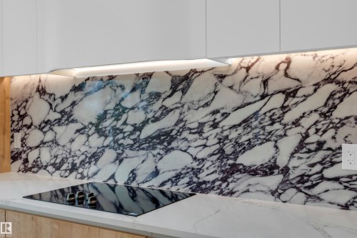 Undercabinet lighting illuminating a polished stone backsplash, white upper cabinetry, light wood-finish lower cabinetry, and a black induction cooktop - 2408 1 Avenue Sw, Edmonton, AB - 