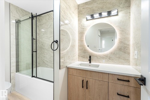 Wood-finish vanity with white countertop, round backlit mirror, matte black faucet, and a tiled shower/tub combination with a clear glass enclosure - 18307 66 Avenue Nw, Edmonton, AB - Indoor Photo Showing Bathroom