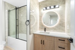 Wood-finish vanity with white countertop, round backlit mirror, matte black faucet, and a tiled shower/tub combination with a clear glass enclosure - 
