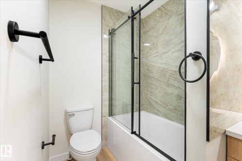 Bathtub with sliding glass shower door, stone-finish shower surround, toilet, and matte black fixtures - 18307 66 Avenue Nw, Edmonton, AB - Indoor Photo Showing Bathroom