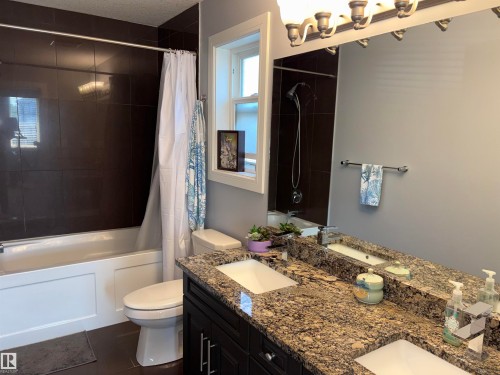 Double vanity with granite countertops and integrated sinks, dark wood-finish cabinetry, full-width mirror with overhead light fixture, and shower-tub combination with dark tile surround - 1452 36A Avenue Nw, Edmonton, AB - Indoor Photo Showing Bathroom