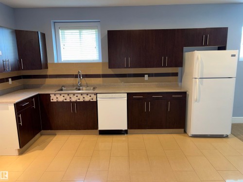 Dark wood-finish cabinetry, light countertops, stainless steel sink with a high-arc faucet, white dishwasher, and light-toned tile flooring - 1452 36A Avenue Nw, Edmonton, AB - Indoor Photo Showing Kitchen With Double Sink