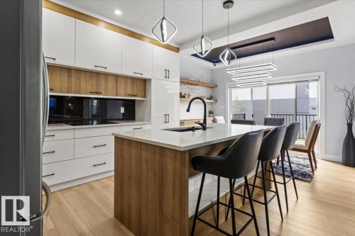 Sleek kitchen featuring white and wood-finish cabinetry, a large kitchen island with a light-toned countertop, wood-finish flooring, and contemporary pendant lighting fixtures - 6903 169 Avenue Nw, Edmonton, AB - Indoor Photo Showing Kitchen