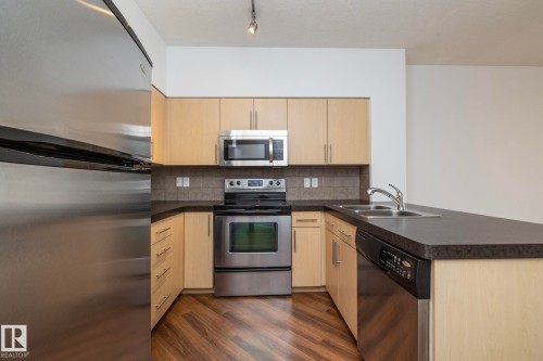 U-shaped kitchen with wood-finish flooring, stainless steel appliances, light wood-finish cabinetry, dark countertops, and a tiled backsplash - #1004 10136 104 St Nw, Edmonton, AB - Indoor Photo Showing Kitchen With Stainless Steel Kitchen With Double Sink