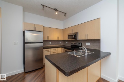 Kitchen featuring stainless steel appliances, light wood-finish cabinetry, dark countertops, a tiled backsplash, and wood-finish flooring - #1004 10136 104 St Nw, Edmonton, AB - Indoor Photo Showing Kitchen With Double Sink