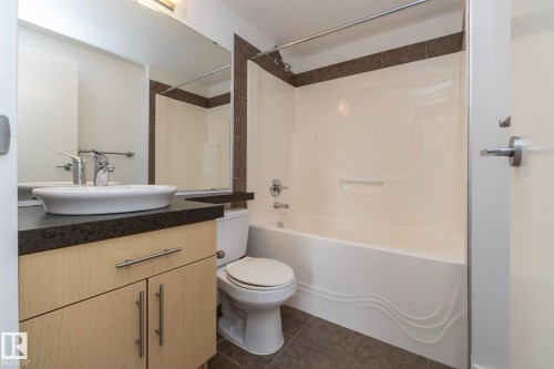 Full vanity mirror, vessel sink, wood-finish cabinetry, dark countertop, and a tiled tub surround - #1004 10136 104 St Nw, Edmonton, AB - Indoor Photo Showing Bathroom