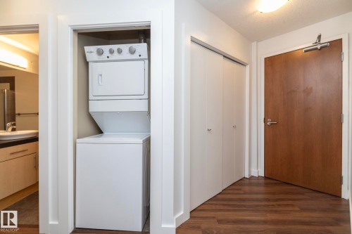 Integrated stacked washer and dryer in a dedicated closet, complemented by wood-finish flooring, a solid core wood entry door, and white bi-fold closet doors - #1004 10136 104 St Nw, Edmonton, AB - Indoor Photo Showing Laundry Room