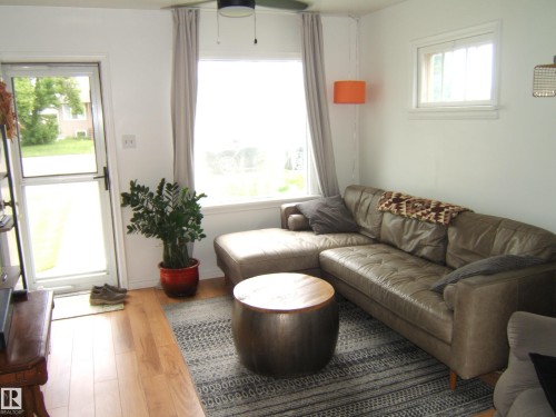 Living area featuring wood-finish flooring, white walls, a large window with drapes, a ceiling fan, and a glass-paneled entry door - 5530 50 Street, Vegreville, AB 
