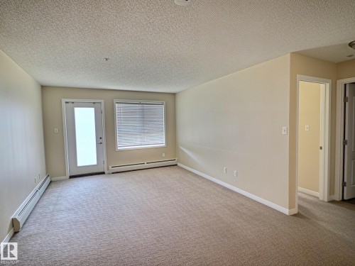 Neutral-toned carpet flooring, textured ceiling, a full-lite exterior door, and a window with horizontal blinds - #111 5350 199 St Nw, Edmonton, AB - Indoor Photo Showing Other Room
