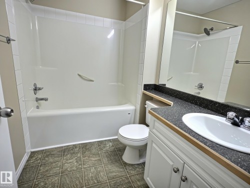 Bathroom featuring a built-in tub and shower combination with a white surround, a vanity with a dark speckled countertop, an undermount sink, and white cabinetry with brushed nickel hardware - #111 5350 199 St Nw, Edmonton, AB - Indoor Photo Showing Bathroom