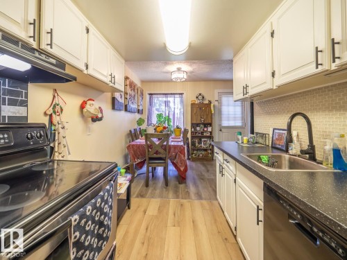 White cabinetry with black hardware, wood-finish flooring, a stainless steel sink with a black pull-down faucet, a tile backsplash, and a built-in electric range - #No 510 Abbottsfield Rd Nw Nw, Edmonton, AB - Indoor Photo Showing Kitchen