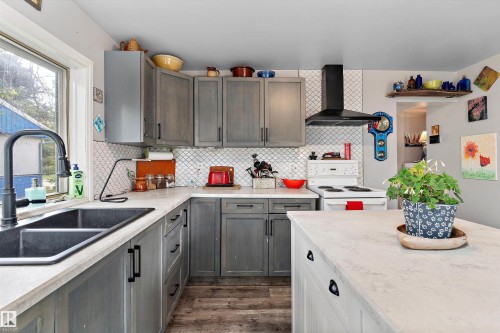 Gray cabinetry with black hardware, white countertop surfaces, white geometric tile backsplash, black dual basin sink, and wood-finish flooring - 5404 45 Avenue, Wetaskiwin, AB - Indoor Photo Showing Kitchen With Double Sink