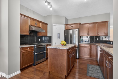 Kitchen featuring wood-finish cabinetry, stainless steel appliances, a kitchen island, granite-style countertops, and a black tile backsplash - 9515 221 Street Nw, Edmonton, AB - Indoor Photo Showing Kitchen With Double Sink