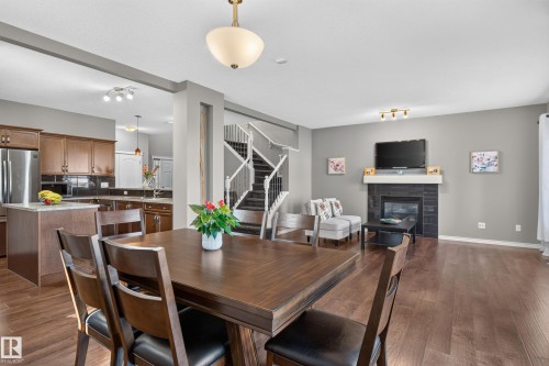 Wood-finish flooring throughout, kitchen with wood cabinetry and stainless-steel refrigerator, central staircase with white railings, living area with fireplace featuring a white mantel, and ceiling-mounted lighting fixtures - 9515 221 Street Nw, Edmonton, AB - Indoor Photo Showing Dining Room With Fireplace