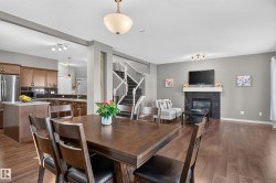Wood-finish flooring throughout, kitchen with wood cabinetry and stainless-steel refrigerator, central staircase with white railings, living area with fireplace featuring a white mantel, and ceiling-mounted lighting fixtures - 