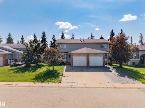 Duplex property featuring a light-colored exterior, two attached garages with white doors and brick accents, individual driveways, and mature trees framing the structure - 4609 38A Avenue Nw, Edmonton, AB - Outdoor With Facade