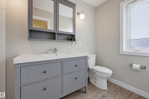 Bathroom featuring a gray vanity with integrated sink, coordinating mirrored medicine cabinet, modern wall sconce, and wood-finish flooring - 12219 93 Street Nw, Edmonton, AB - Indoor Photo Showing Bathroom