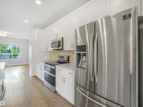 Stainless steel French door refrigerator, white shaker-style cabinetry, wood-finish flooring, recessed lighting, and a window with green foliage beyond - 203 Cavanagh Common Sw, Edmonton, AB - Indoor Photo Showing Kitchen With Upgraded Kitchen