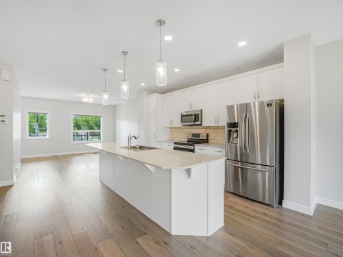 Open-concept kitchen featuring an island with sink, white shaker cabinets, stainless steel appliances, and light wood-finish flooring - 203 Cavanagh Common Sw, Edmonton, AB - Indoor Photo Showing Kitchen With Upgraded Kitchen