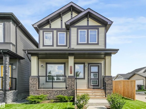 Two-story residence featuring light-colored siding, a covered front porch with brick and glass railing, a dark front door with window panels, and multiple windows with dark trim - 203 Cavanagh Common Sw, Edmonton, AB - Outdoor With Deck Patio Veranda