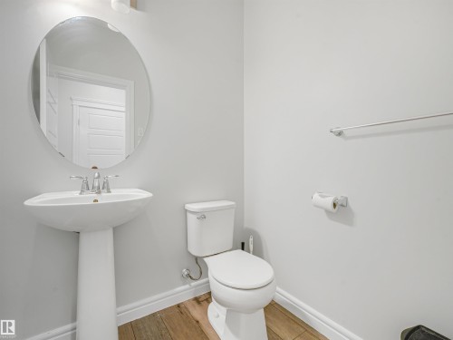 Pedestal sink with chrome faucet, oval mirror, toilet, and wood-finish flooring - 203 Cavanagh Common Sw, Edmonton, AB - Indoor Photo Showing Bathroom