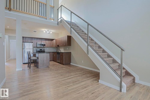 Open-concept living area with wood-finish flooring, a kitchen island, stainless steel appliances, and a glass-paneled staircase with carpeted treads - #607 10518 113 St Nw Nw, Edmonton, AB - Indoor Photo Showing Other Room