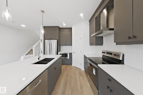 Modern kitchen featuring a large island with an undermount sink and chrome faucet, stainless steel appliances, grey cabinetry, white countertops, and light wood-finish flooring - 7791 Eifert Crescent Nw, Edmonton, AB - Indoor Photo Showing Kitchen With Double Sink With Upgraded Kitchen