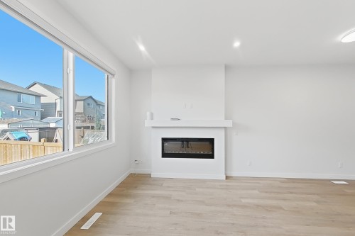 Living space featuring an integrated electric fireplace with a mantel, light wood-finish flooring, white walls, and large windows - 7791 Eifert Crescent Nw, Edmonton, AB - Indoor Photo Showing Other Room With Fireplace