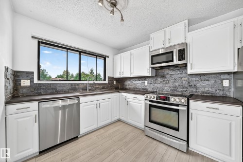Kitchen featuring white cabinetry, stainless steel appliances, a stacked stone-look backsplash, wood-finish flooring, and a large window - 16808 100 Street Nw, Edmonton, AB - Indoor Photo Showing Kitchen With Stainless Steel Kitchen With Upgraded Kitchen