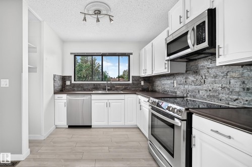Kitchen featuring white cabinetry, stainless steel appliances, dark countertops, a stone-finish backsplash, and wood-finish flooring - 16808 100 Street Nw, Edmonton, AB - Indoor Photo Showing Kitchen With Stainless Steel Kitchen With Upgraded Kitchen