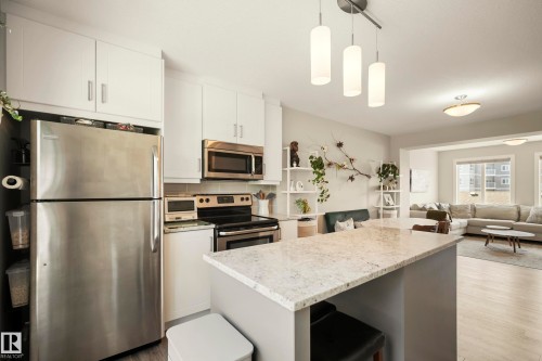 Modern kitchen and great room featuring stainless steel appliances, white cabinetry with ample storage, quartz-finish island countertop with seating, light wood-finish flooring, pendant lighting, and open sightlines to the living area with large windows - 4013 Blackbird Link Nw, Edmonton, AB - Indoor Photo Showing Kitchen