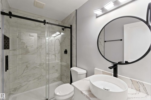 Bathroom featuring a glass-enclosed shower with black hardware, a shower niche with honeycomb tile, a floating vanity with a vessel sink, a black-framed circular mirror, and a wall-mounted light fixture - 4129 134A Avenue Nw, Edmonton, AB - Indoor Photo Showing Bathroom