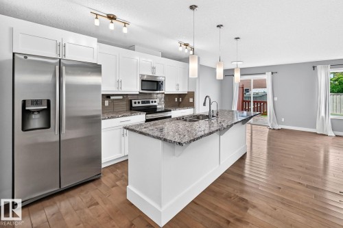 White cabinetry, stainless steel appliances, wood-finish flooring, kitchen island with dual sinks, and pendant lighting - 1112 176 Street Sw, Edmonton, AB - Indoor Photo Showing Kitchen With Double Sink With Upgraded Kitchen