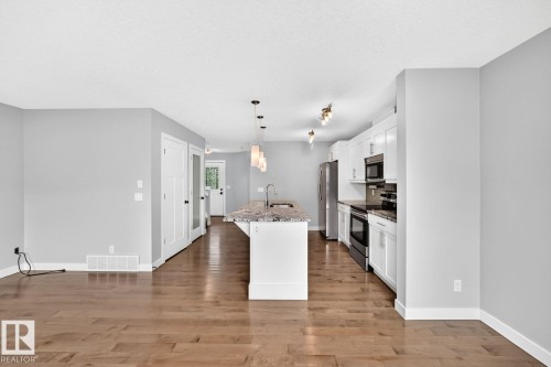 Open-concept living space featuring wood-finish flooring, a kitchen island with pendant lighting, white cabinetry, and stainless steel appliances - 1112 176 Street Sw, Edmonton, AB - Indoor Photo Showing Kitchen