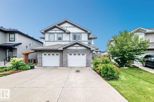Two-story residence featuring light gray siding with dark gray trim, stone veneer accents, double attached garage with white doors, concrete driveway, and landscaped front yard with mature shrubbery and a tree - 1112 176 Street Sw, Edmonton, AB - Outdoor With Facade