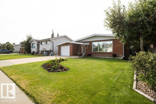 Brick exterior with white trim, attached garage, concrete driveway, front yard with mature landscaping, and an additional landscaped garden bed - 10729 108A Avenue, Westlock, AB - Outdoor