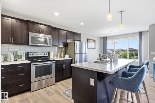 Kitchen featuring dark wood-finish cabinetry, stainless steel appliances, light-toned countertops, and a kitchen island with integrated sink - 944 Morris Way, Leduc, AB - Indoor Photo Showing Kitchen With Double Sink With Upgraded Kitchen