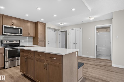 Open-concept kitchen featuring warm wood-finish cabinetry, stainless steel appliances, white subway tile backsplash, and a spacious island with quartz countertop - #131 5504 Schonsee Dr Nw, Edmonton, AB - Indoor Photo Showing Kitchen