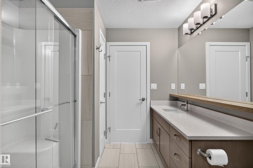 Vanity with dual sinks and light-toned countertop, large mirrored wall, shower with glass enclosure, wood-look tile flooring - #131 5504 Schonsee Dr Nw, Edmonton, AB - Indoor Photo Showing Bathroom