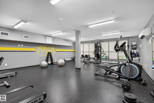 Fitness center with rubber flooring, fluorescent lighting, and large windows with roller shades - #131 5504 Schonsee Dr Nw, Edmonton, AB - Indoor Photo Showing Gym Room