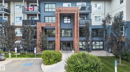 Multi-story building featuring a brick and siding facade, multiple balconies with black railings, and a prominent entrance - #131 5504 Schonsee Dr Nw, Edmonton, AB - Outdoor With Balcony With Facade