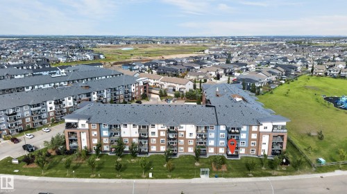 Multi-story building with a dark gray roof, featuring multiple balconies, and a landscaped green space immediately surrounding the structure - #131 5504 Schonsee Dr Nw, Edmonton, AB - Outdoor With View