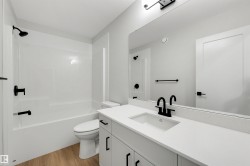 White vanity with integrated sink, white countertop, matte black fixtures, white bathtub with shower, and wood-finish flooring - 